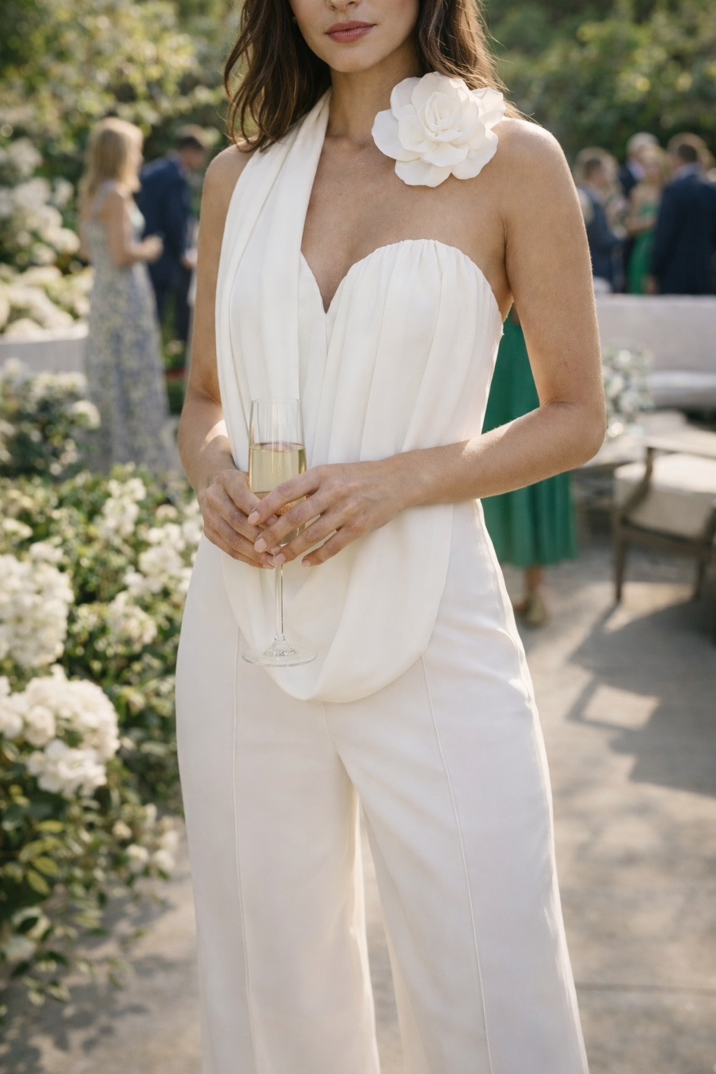 Model wearing an elegant white halter-neck jumpsuit with a draped front, wide-leg silhouette, and floral shoulder detail at an outdoor garden event.