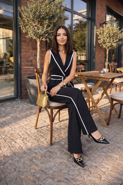 Woman styled in a black sleeveless wrap jumpsuit with white contrast trim and a tied waist belt, holding a gold purse on an outdoor patio.